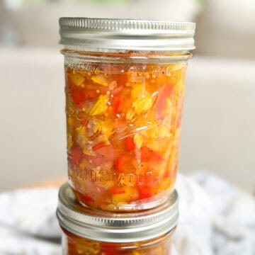 A close-up image of two glass mason jars filled with a colorful mixture of chopped vegetables, likely a homemade relish or salsa. The jars are sealed with metal lids and the top jar is slightly angled, resting on a blurry background.
