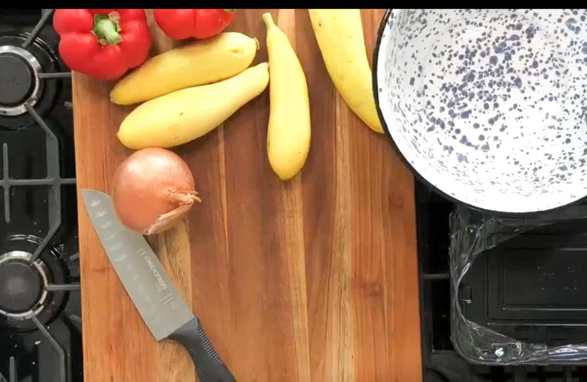 A bamboo cutting board holds a knife, three yellow squashes, a red bell pepper, and an onion. A speckled white bowl and a stove are visible adjacent to the board.