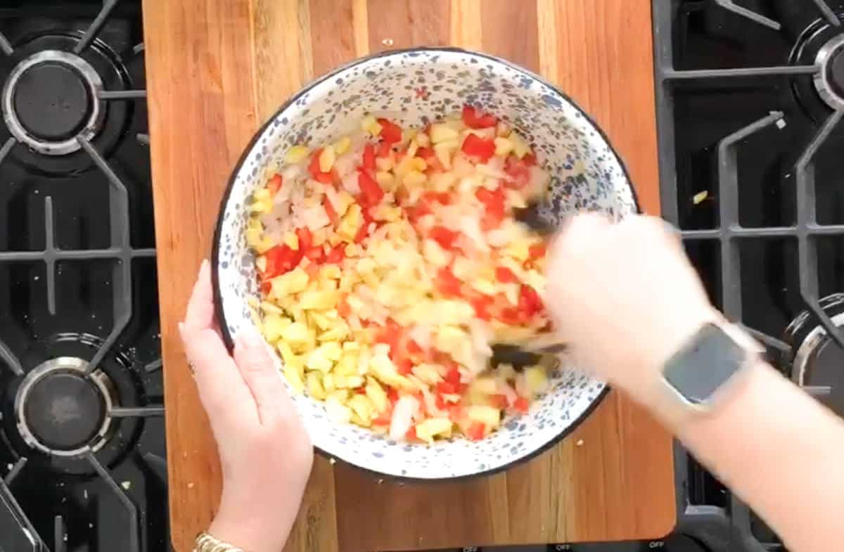 A person is stirring a mixture of chopped vegetables in a white bowl with a floral pattern. The bowl is placed on a wooden cutting board between two stove burners. The person's watch is visible on their wrist.