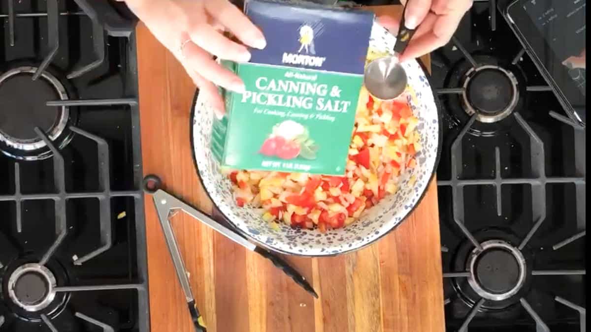 A pair of hands is holding a box of Morton Canning & Pickling Salt above a bowl filled with chopped vegetables. The bowl is on a wooden cutting board situated between two stove burners. A measuring spoon and a pair of scissors are also on the board.