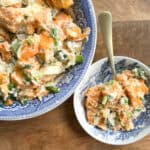 A large blue and white bowl and a smaller matching bowl contain servings of a salad made with sweet potatoes and garnished with green onions. A gold-colored spoon rests in the smaller bowl. Both bowls sit on a wooden surface.
