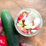 A top view of a glass jar filled with sliced cucumbers, tomatoes, and onions. Surrounding the jar on a wooden surface are a cucumber, red onion, and two red bell peppers.