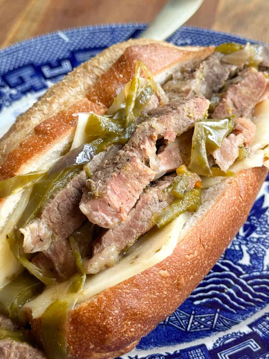 Close-up view of a beef sandwich on a crusty roll, topped with slices of cooked beef, melted cheese, and green bell peppers. The sandwich is served on a blue and white patterned plate.