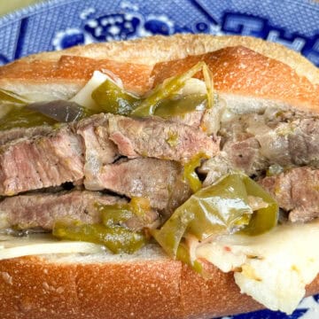 Close-up of an Italian beef sandwich with slices of beef and green peppers, served on a French roll. Plate with a blue and white design in the background.