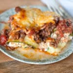 A close-up of a serving of zucchini lasagna on a green plate. The lasagna features layers of zucchini, meat sauce, ricotta cheese, and melted cheese on top, with some sauce and cheese spilling over the side. A fork is partially visible in the background.