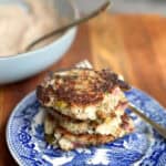 A close-up of a stack of three hash brown patties on a blue and white plate. The hash browns appear golden brown and crispy. There is a fork resting on the plate. A bowl of sauce with a spoon in the background is slightly out of focus.
