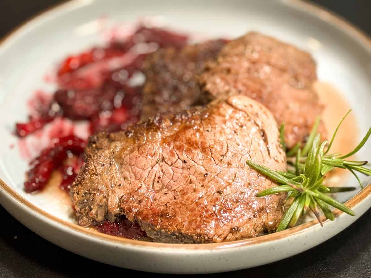 A plate of cooked meat, seasoned and garnished with a sprig of rosemary. Accompanied by a red sauce or compote, with both the meat and sauce presented on a white plate with a light-colored rim.