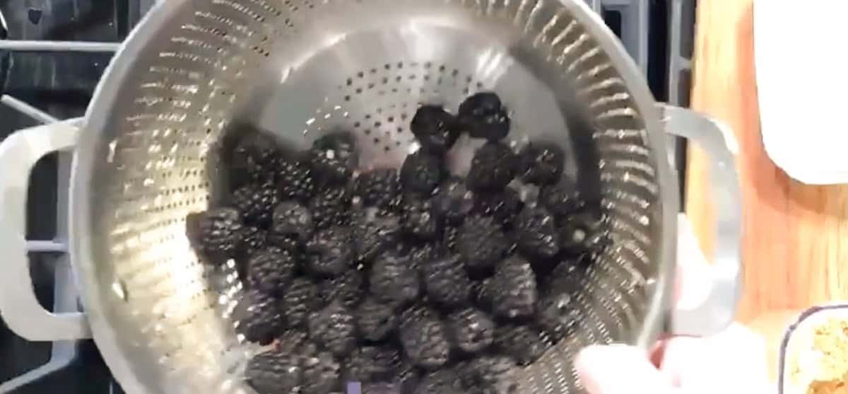 A person holds a colander filled with washed blackberries over a stove. The colander is silver with handles on both sides, and the blackberries are dark and ripe. Part of a wooden cutting board is visible on the side.