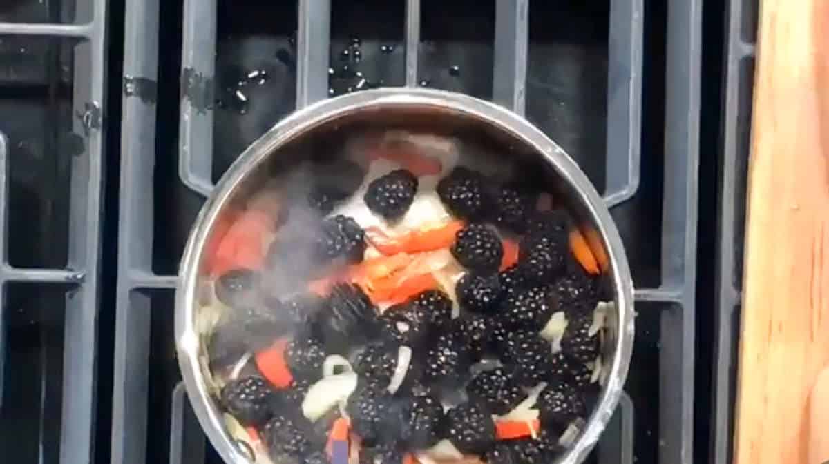 A saucepan on a stovetop grill contains a mixture of blackberries, sliced red bell peppers, and onions, with steam rising from the food.