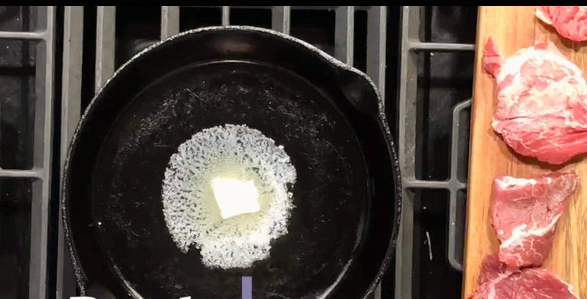 A cast-iron skillet on a stovetop with a piece of butter melting in the center. To the right, a wooden cutting board holds several raw meat slices.