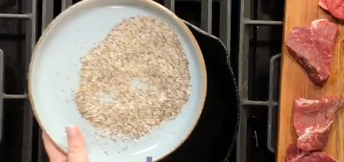 A hand places a light blue plate with a mix of spices onto a black surface. To the right on a wooden cutting board are raw steak pieces, and the background appears to be a stove with a cast iron skillet visible.
