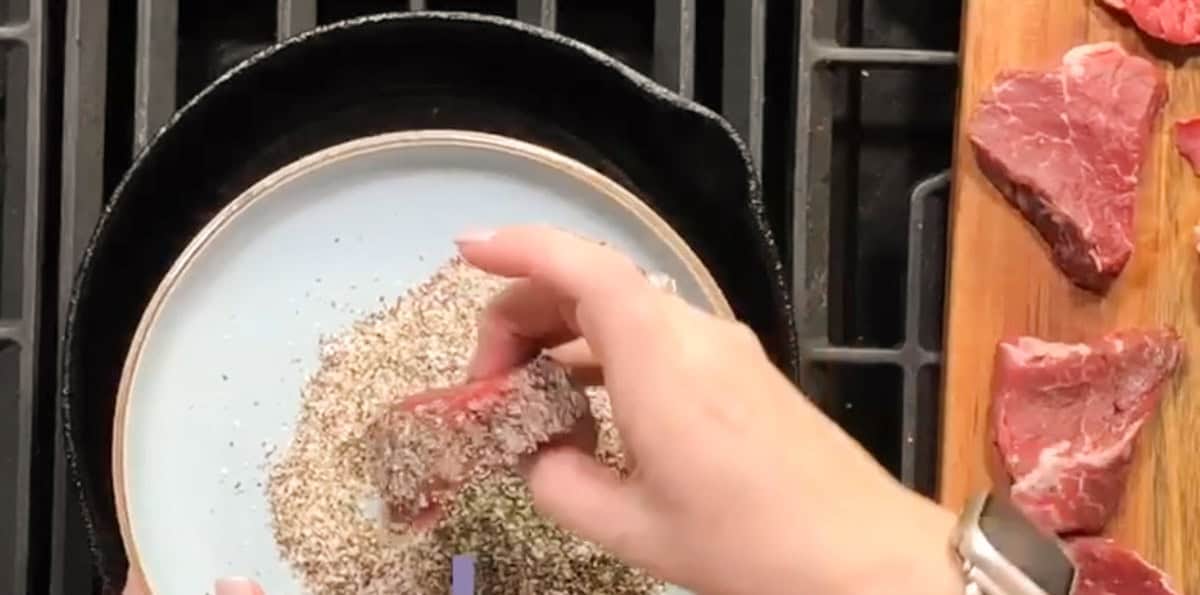 A person is dipping a piece of raw steak into a plate filled with pepper and seasoning. The steak is partially coated with the seasoning. Several other pieces of raw steak can be seen on a wooden cutting board to the right. This is occurring over a stovetop.