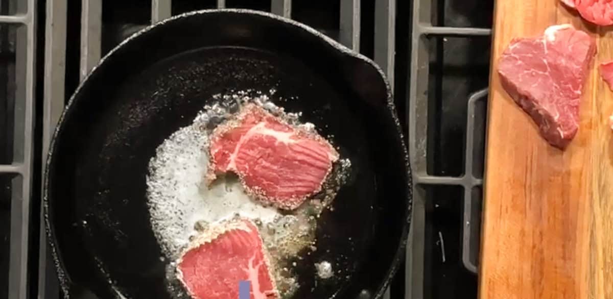 A black skillet on a stove contains two pieces of raw meat being cooked in butter. To the right, a wooden cutting board holds another piece of raw meat and a small piece of parsley.