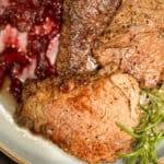 Close-up of a plate with cooked beefsteak garnished with spices. To the side, there are pieces of a red, chunky sauce, and a sprig of rosemary. The meat appears well-seasoned and seared.
