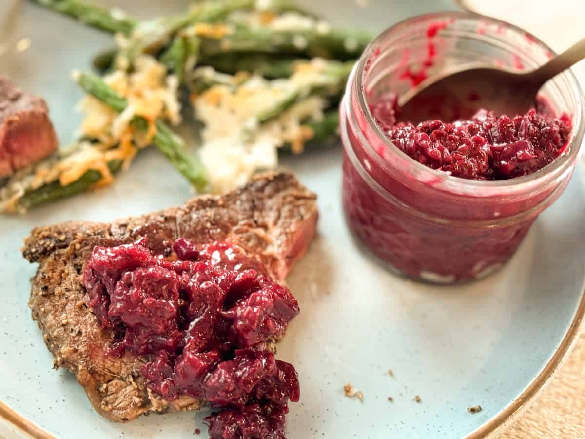 A dish with a piece of cooked meat topped with a chunky red sauce, a side of green beans with melted cheese, and an open jar of the same red sauce with a spoon inside, all on a light blue plate.