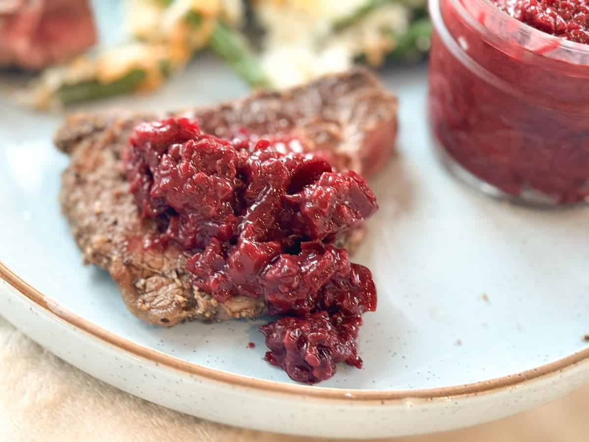 A close-up view of a steak topped with a chunky red sauce, served on a light blue plate. In the background, there are some green vegetables and what appears to be more of the red sauce in a small container.
