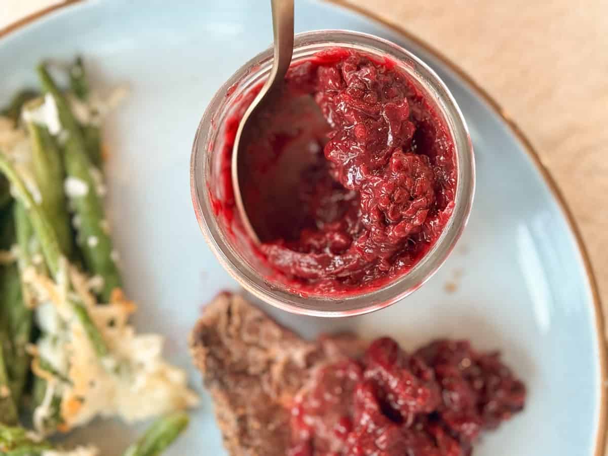 A jar of red raspberry jam with a spoon inside sits on a white plate. Next to the jar are green beans topped with cheese and a piece of meat with jam spread on top. The image captures a top-down view of a meal.