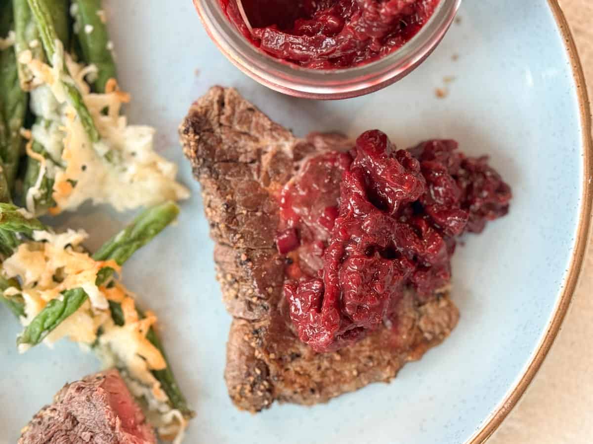 A plate with a portion of steak topped with a cranberry sauce, green beans with a creamy white sauce and cheese, and a small jar containing additional cranberry sauce is placed on a light blue ceramic plate.