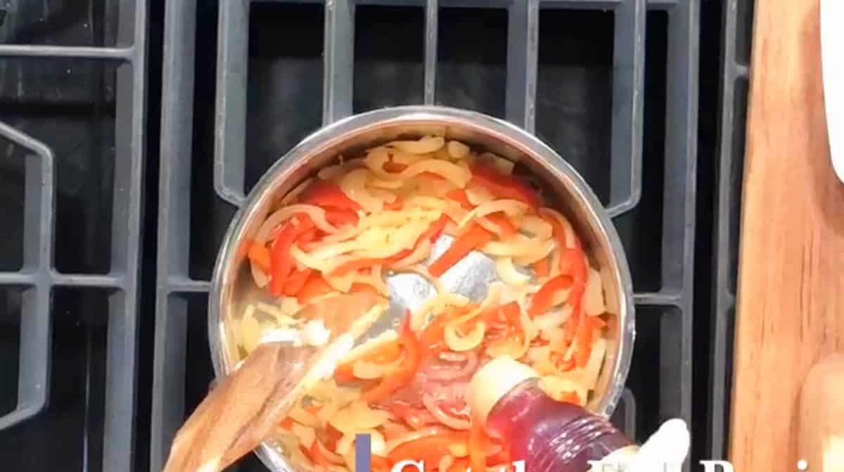 A top-view photo of a stainless steel pot on a stove, with sliced bell peppers and onions being cooked. A hand pours a liquid from a bottle into the pot. Two wooden utensils are seen stirring the contents.