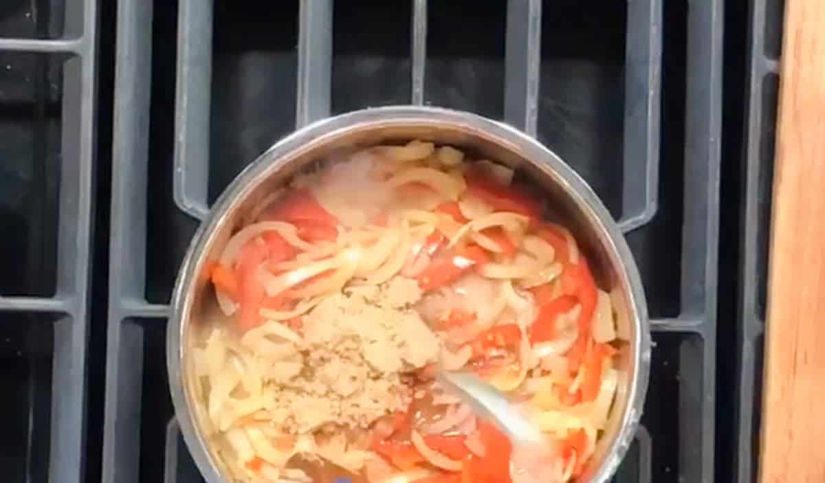 A stainless steel pot on a stove contains sliced onions and red bell peppers. There is a mound of brown sugar in the middle of the mixture, and a spoon is partially visible on the right side of the pot, stirring or resting on the mixture.