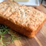 A freshly baked loaf of bread sits on a wooden cutting board. The bread has a golden-brown crust and a rustic appearance. Some sprigs of rosemary are placed next to the loaf. A plate with a stick of butter is visible in the background.