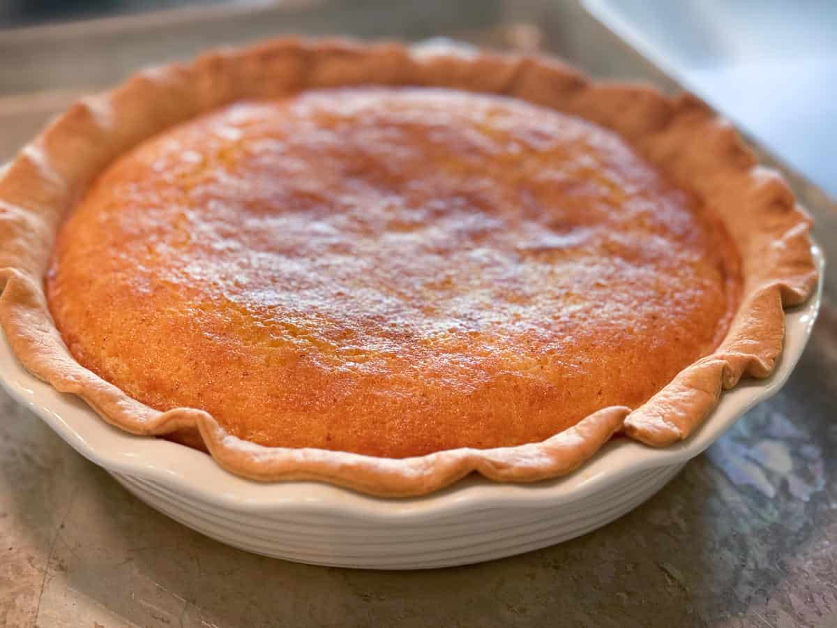 A close-up view of a freshly baked pie with a golden-brown top and crimped crust edges, placed in a white ceramic pie dish on a light-colored surface.