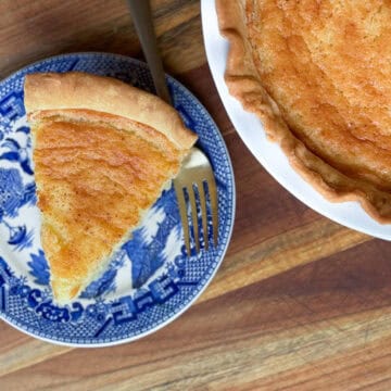 A slice of pie on a blue and white plate next to a fork. The plate is beside a pie in a white dish, both placed on a wooden surface. The pie has a golden brown crust and filling.
