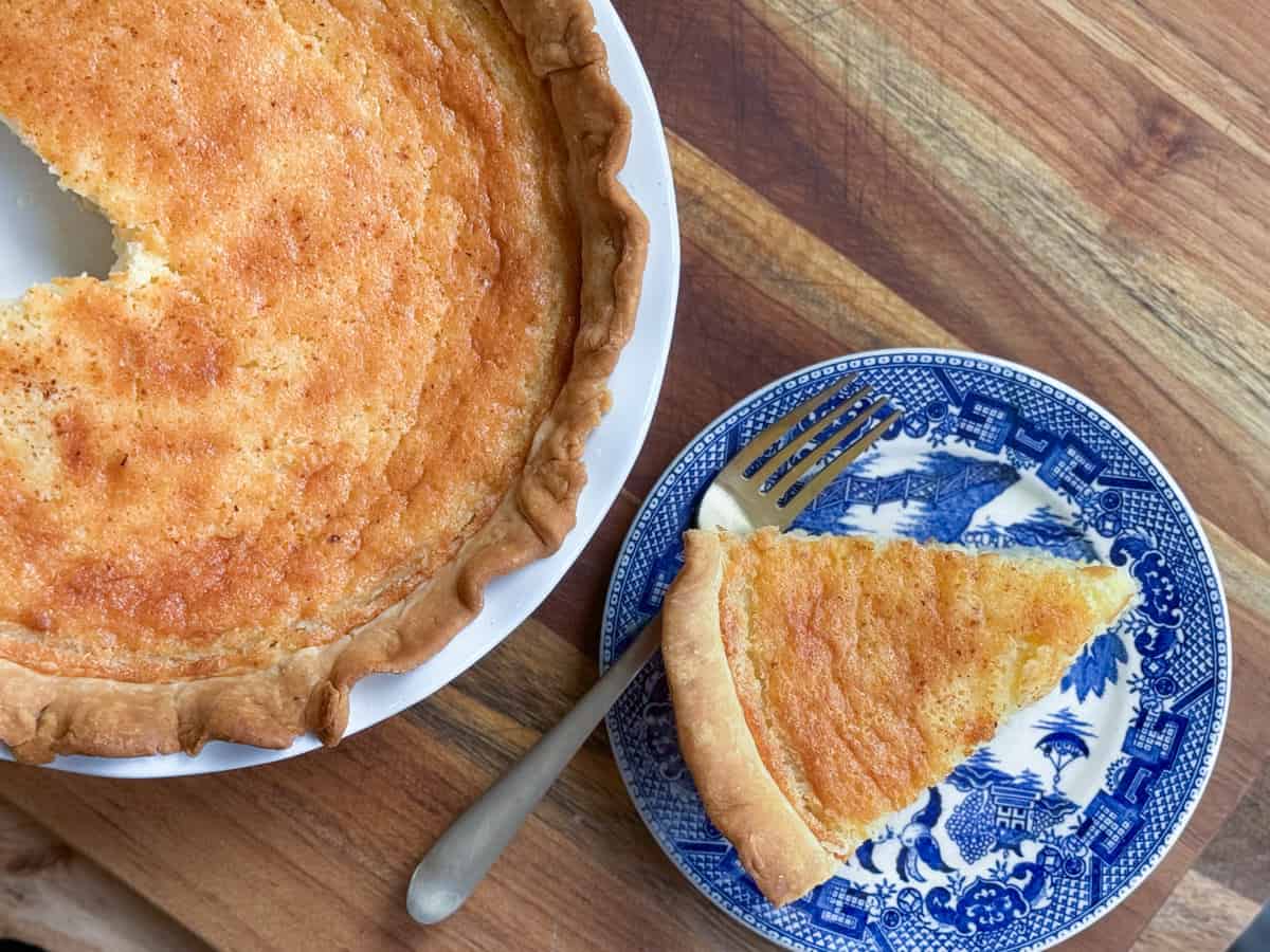 A whole custard pie with a slice missing sits on a wooden surface. The missing slice is placed next to it on a blue and white patterned plate, along with a fork. The pie has a golden-brown crust and a firm, slightly browned filling.
