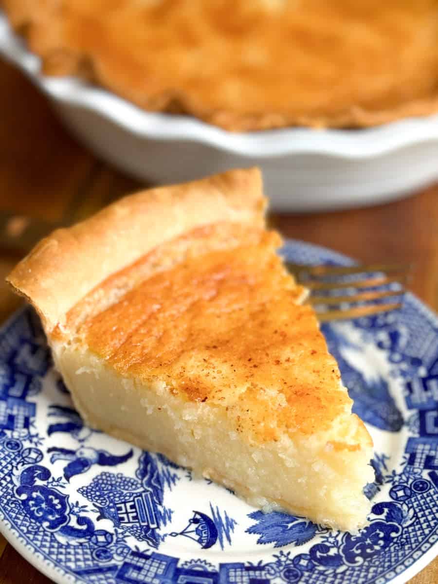 A slice of custard pie on a blue and white patterned plate, with a fork resting beside it. The pie has a golden-brown top and a creamy texture. The rest of the pie is visible in a white dish in the background.