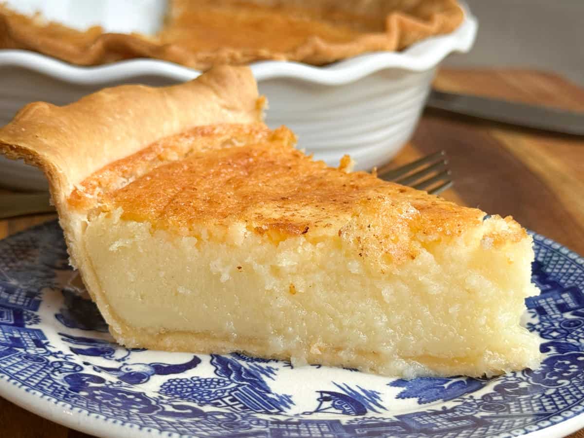 A close-up of a slice of custard pie on a decorative blue and white plate. The pie has a golden brown crust and a smooth, creamy filling. A fork is partially visible in the background next to a white pie dish.