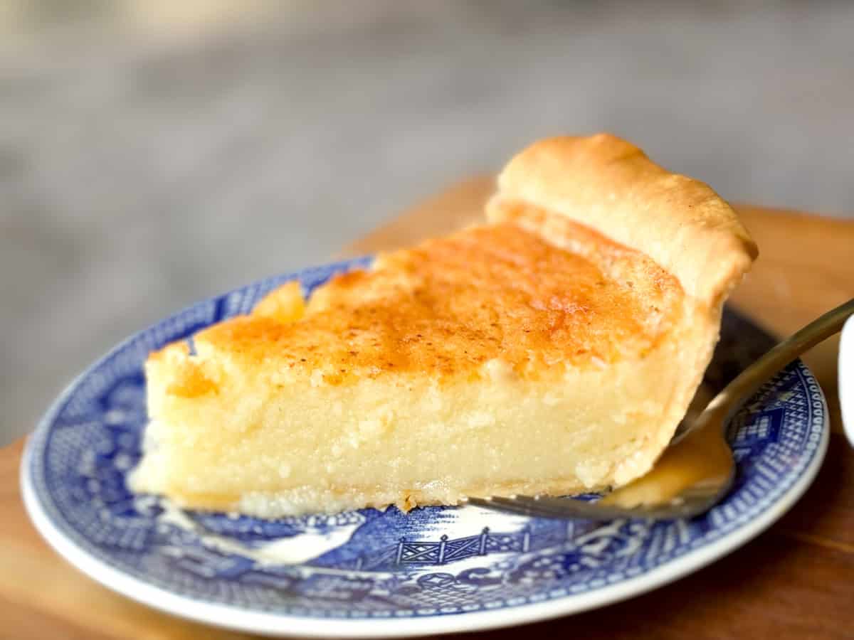 A slice of custard pie with a golden brown top is served on a blue and white patterned plate. The creamy filling appears smooth and slightly firm, and a fork lies next to the slice on the plate. The background is blurred.