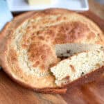 A round, golden-brown Irish soda bread with a crispy crust sits on a wooden surface. A slice has been cut from the loaf, showing a slightly moist interior with bits of dried fruit. A plate with a stick of butter is in the background.