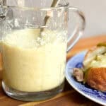 A glass measuring cup filled with a creamy liquid sits on a wooden surface. A metal spoon is placed inside the measuring cup. Beside it, on the right, is a plate with a sandwich on a blue and white patterned plate.