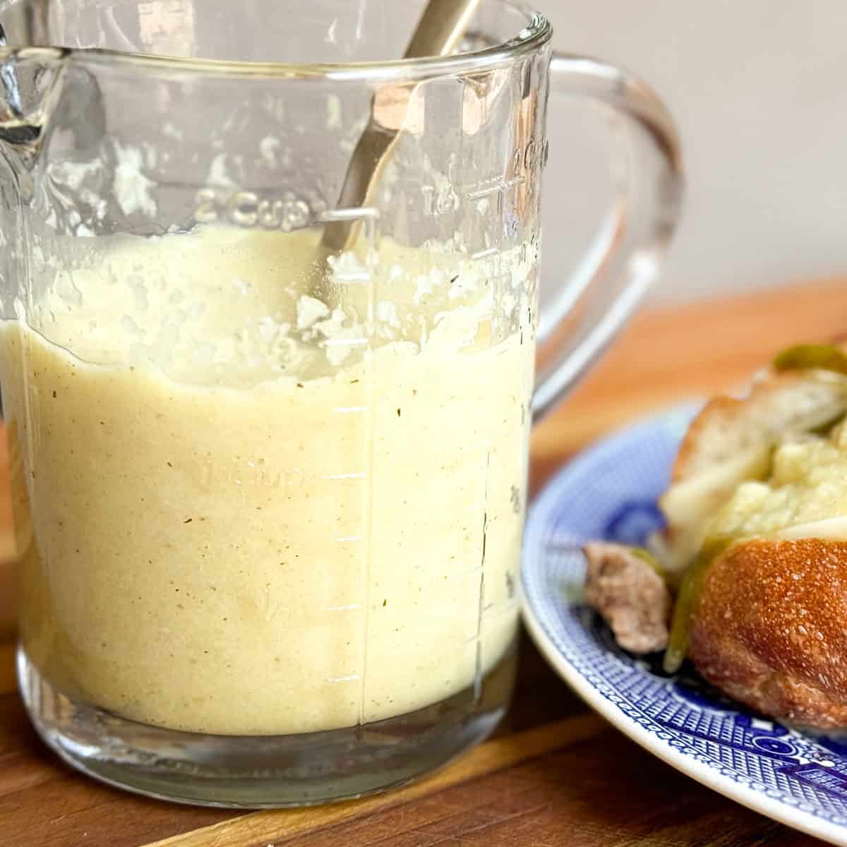 A glass measuring cup filled with a creamy liquid sits on a wooden surface. A metal spoon is placed inside the measuring cup. Beside it, on the right, is a plate with a sandwich on a blue and white patterned plate.