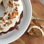 A partially eaten, frosted bundt cake on a white plate. The cake is topped with chopped nuts and orange zest. A small slice is placed on the wooden surface next to the plate, also showing its frosting and toppings. A fork is nearby.