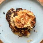A close-up of three cooked almond flour Johnny cakes stacked on a light blue plate. The cakes are slightly overcooked with a golden brown and dark brown crust, and crumbs are scattered on the plate around them.