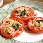 Three slices of tomato topped with melted cheese, basil leaves, and other seasonings are arranged on a white plate with a green pattern. The tomatoes appear to be baked or grilled. The background includes part of a table surface.