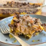 A portion of a casserole made with ground beef, melted cheese, and another ingredient, possibly potatoes or pasta, is served on a plate with a fork. The baking dish containing the remaining casserole is visible in the background.