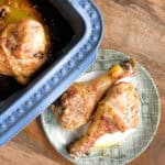Two roasted chicken drumsticks placed on a round, green-patterned plate next to a blue baking dish containing another drumstick. The food is set on a wooden surface.