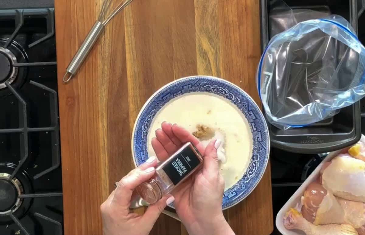 A person is holding a small bottle of ground ginger, sprinkling some into a bowl with a creamy mixture. The bowl is on a wooden countertop, near a whisk, a stovetop, and a container with raw chicken pieces. There is also a plastic bag next to the chicken.