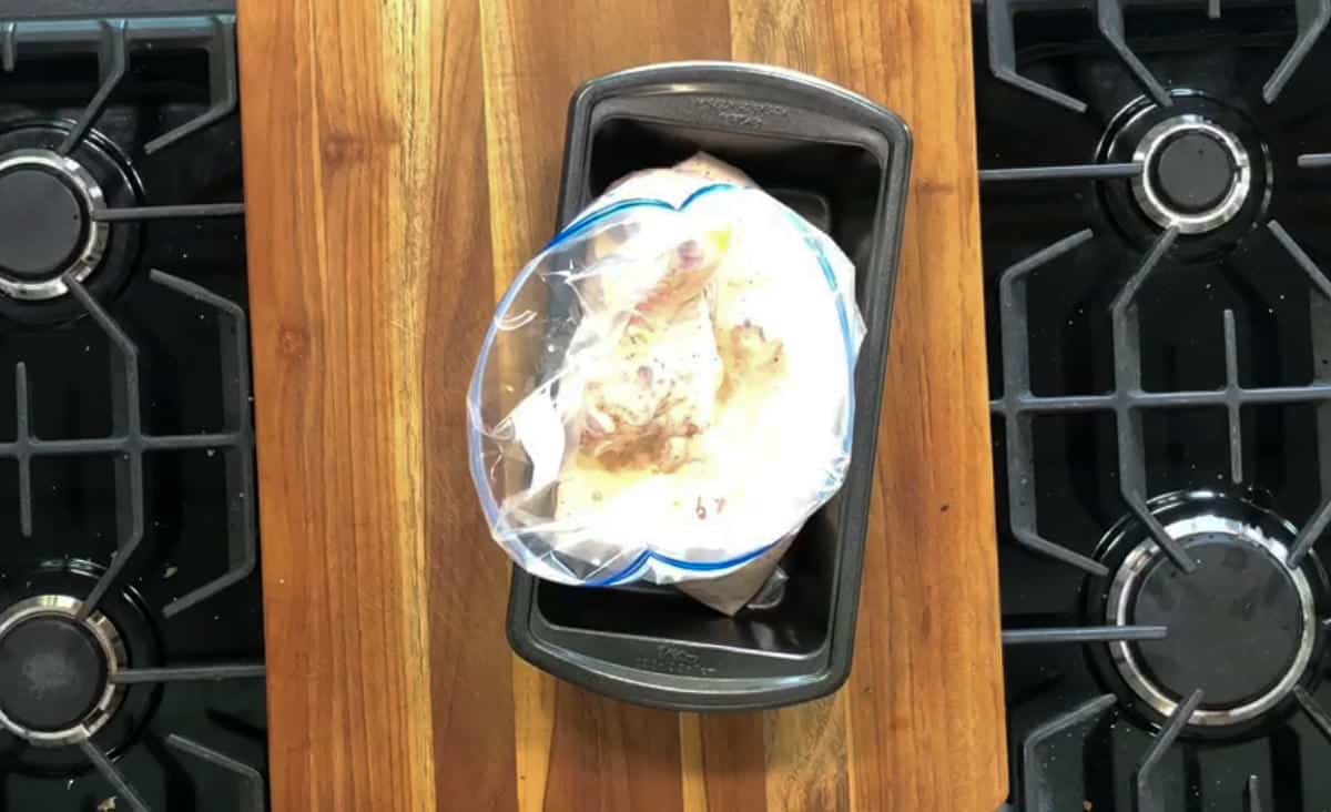 A plastic bag containing marinated chicken is placed inside a black rectangular baking dish. The baking dish rests on a wooden cutting board in the center of a stovetop with two burners visible on either side.