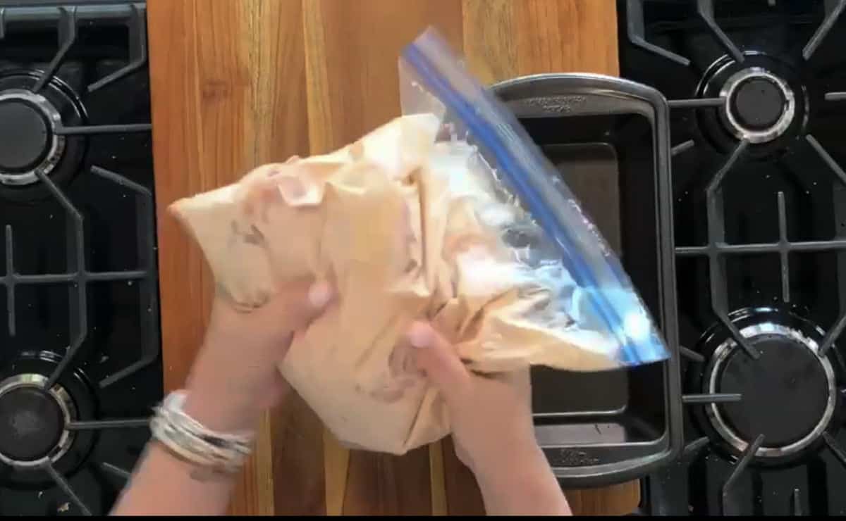 Two hands are holding a sealed plastic bag containing marinated chicken pieces over a wooden surface. Two stove burners and a baking tray are visible on either side.