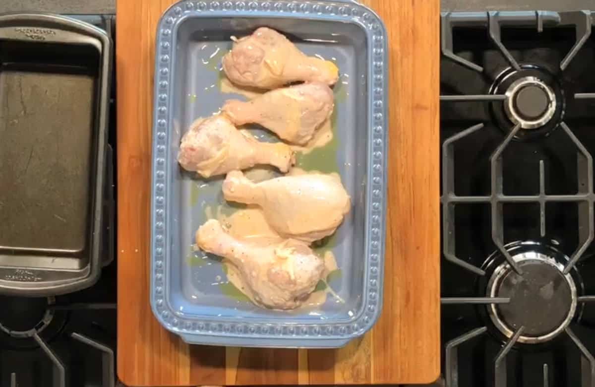 A blue rectangular dish with five uncooked, marinated chicken drumsticks is placed on a wooden cutting board resting on a stovetop. Next to it is a small baking pan. The stove burners are visible on the right side.