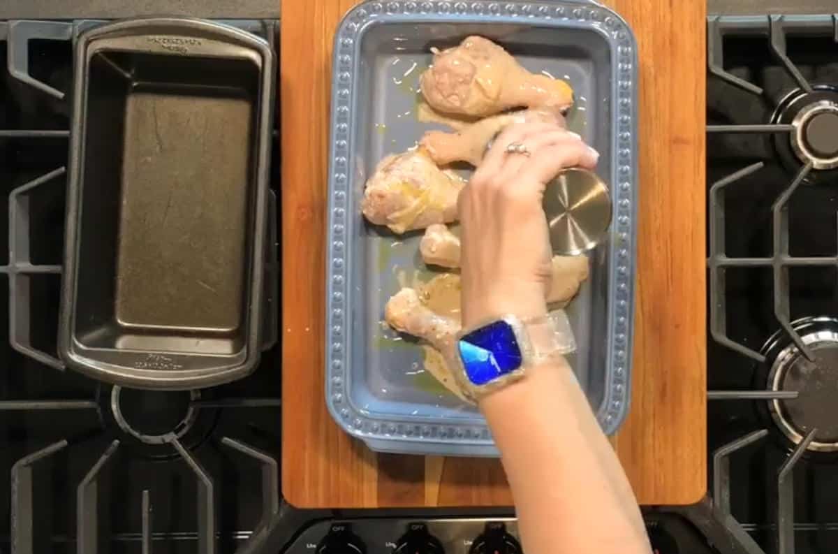 A person's hand is seasoning raw chicken drumsticks in a blue baking dish placed on a wooden board over a stove. A loaf pan is on the left side of the dish. The person is wearing a watch with a blue screen.