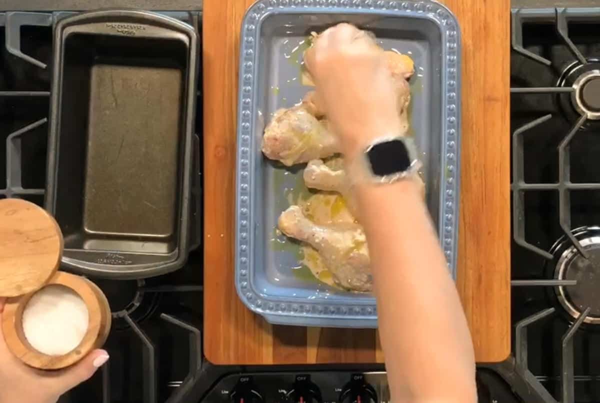 A person is seasoning chicken drumsticks with salt in a blue baking dish on top of a wooden cutting board. The dish is placed on a stove. A loaf pan and a salt container are next to the dish. The person is wearing a smartwatch on their wrist.