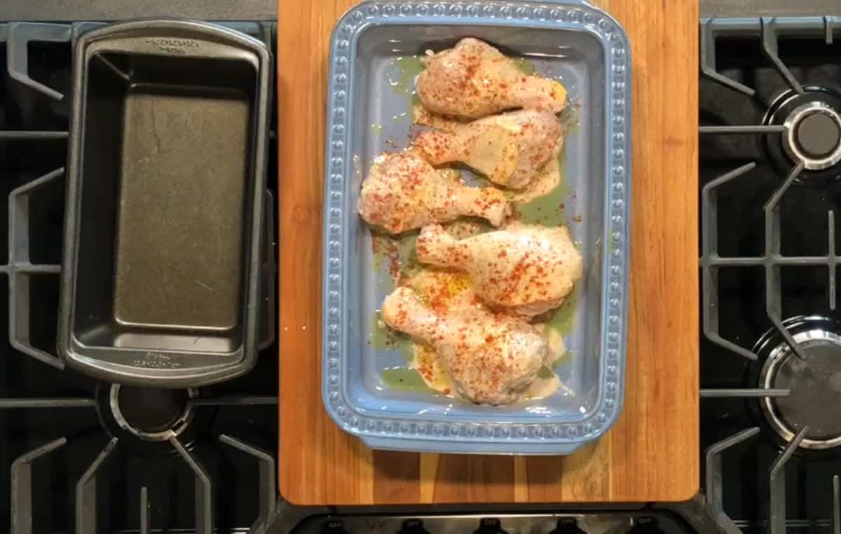 A ceramic dish containing uncooked, seasoned chicken drumsticks is placed on a wooden cutting board. The cutting board is situated on a gas stove with a black loaf pan placed on the left side.
