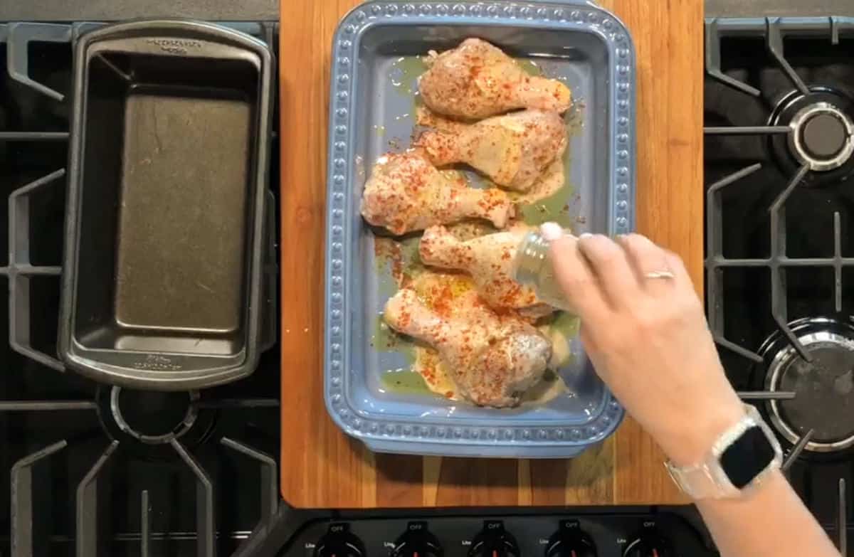 A person's hand is sprinkling seasoning onto four raw chicken drumsticks in a blue baking dish. The dish is placed on a stovetop, and a loaf pan sits to the left on a wooden cutting board. The person is wearing a watch.