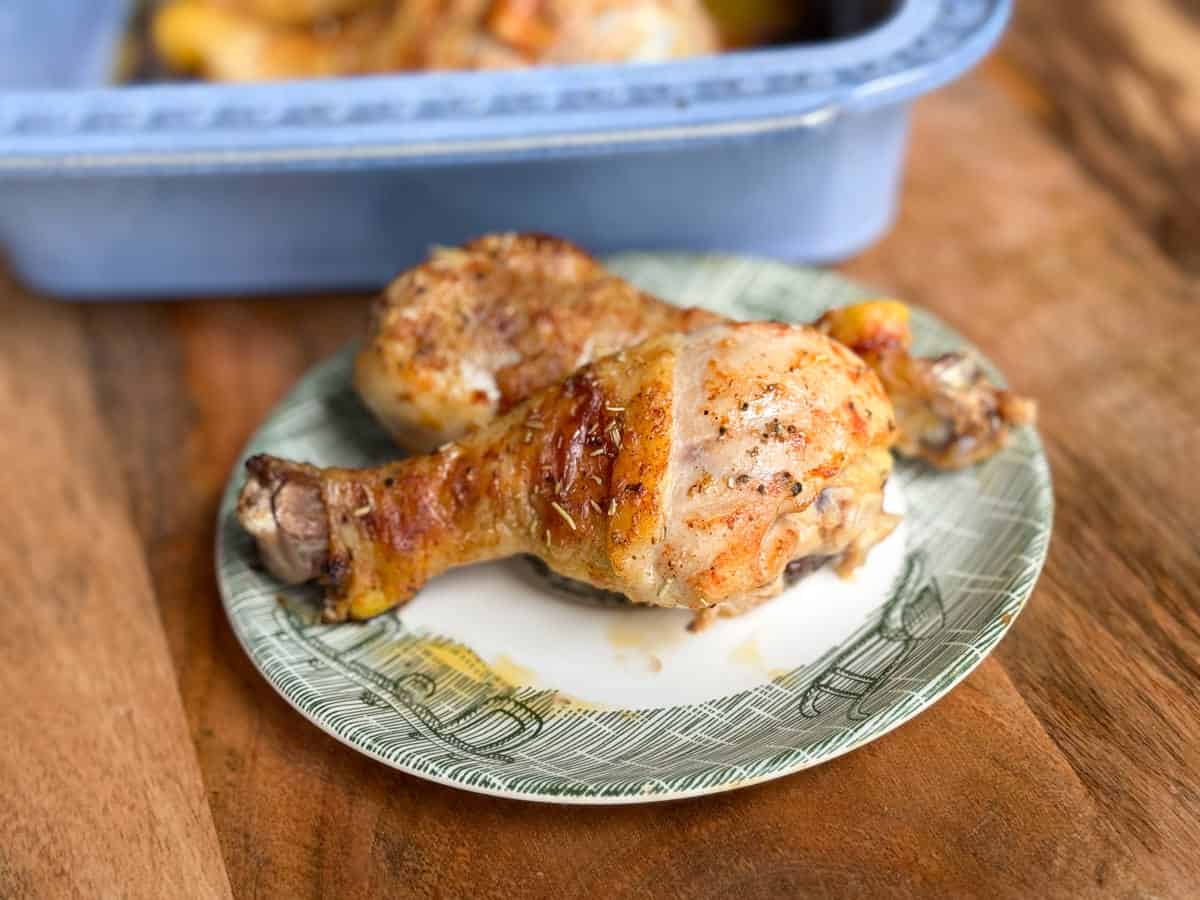 A close-up of a plate with a cooked chicken drumstick and a piece of roasted chicken thigh on a wooden table. A blue baking dish with more chicken is in the background.