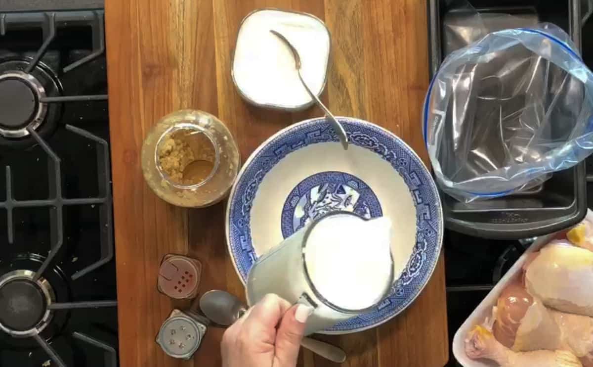 A hand holding a measuring cup filled with white liquid is pouring it into a bowl with blue decorative patterns. Various cooking ingredients and tools, including spices, sugar, a jar of mustard, and raw chicken pieces in a plastic bag, are on the wooden surface.