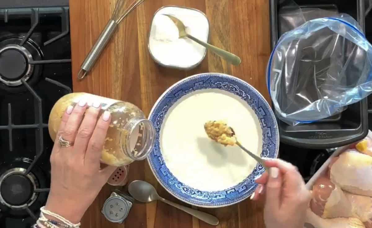 Top-down view of a kitchen setup showing hands preparing a mixture in a blue and white bowl on a wooden countertop. Ingredients include a jar of minced garlic, a container of white powder, and uncooked chicken pieces in a plastic-lined container.